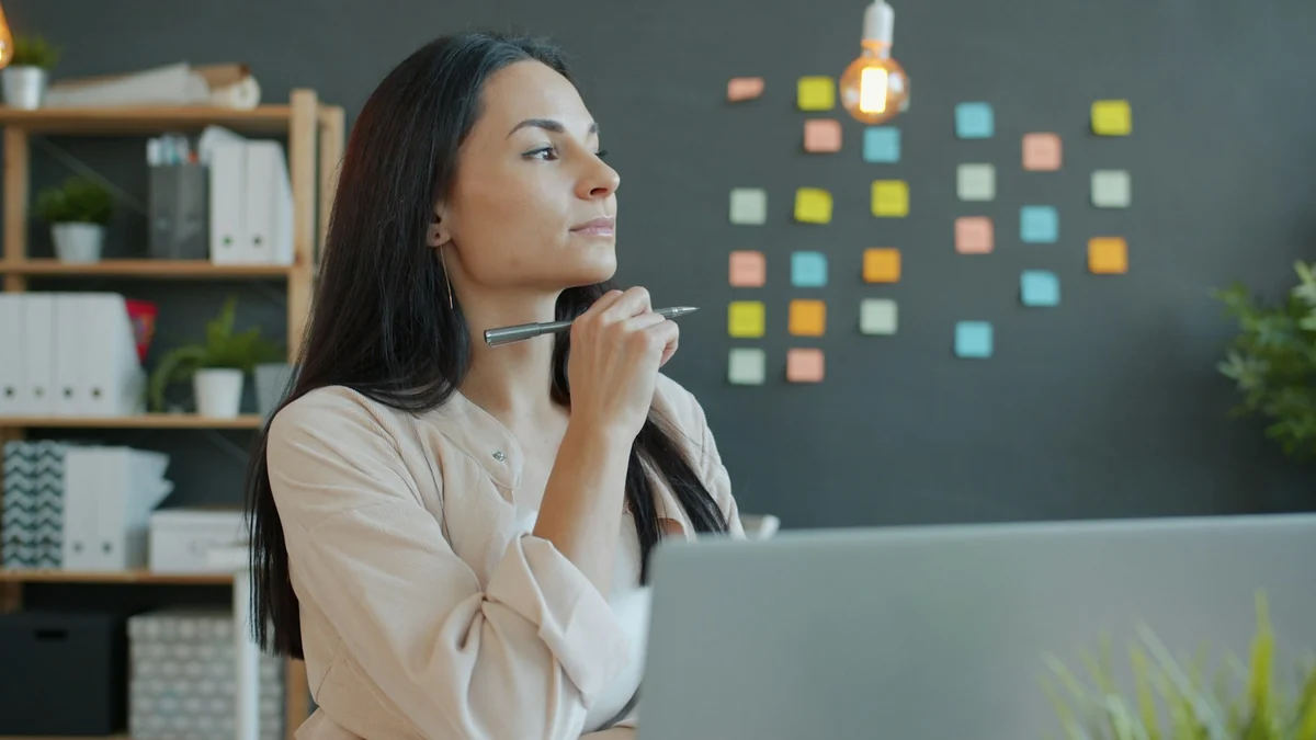 A woman holding a pen and thinking in front of a laptop, with a sticky note board behind her