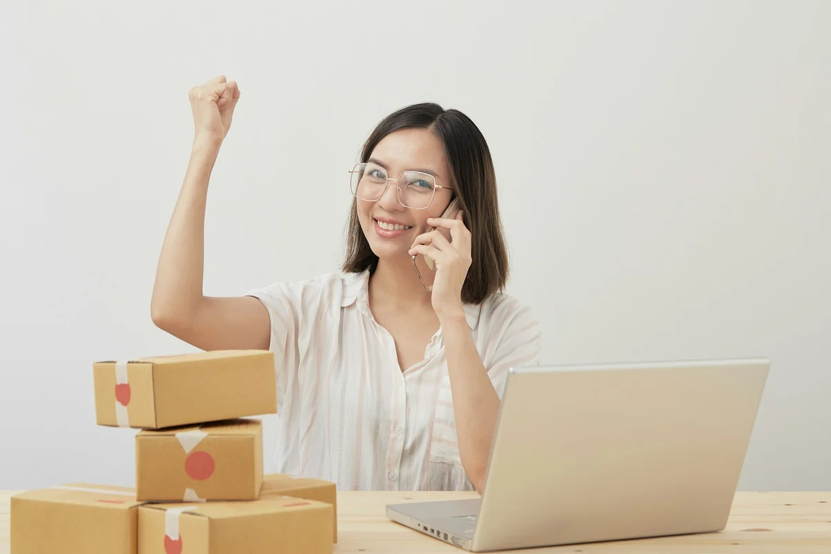 Shop owner in glasses on the phone next to a laptop and a stack of packed boxes
