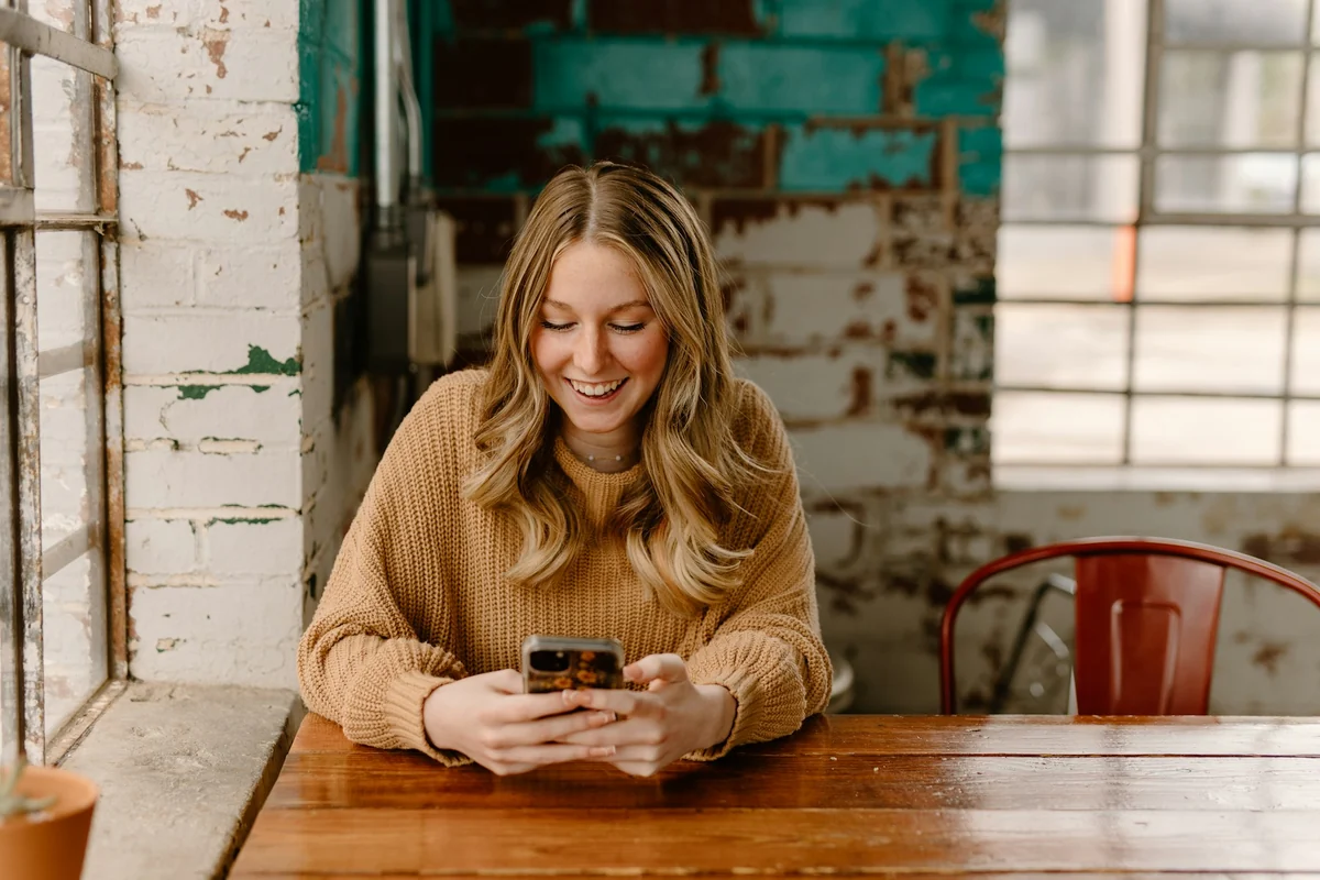 A woman at a wooden table smiling as she types a message on her phone