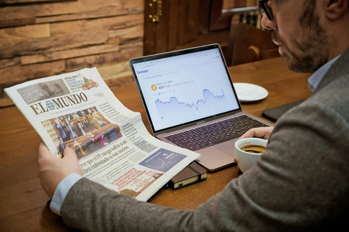 A man reading a printed newspaper next to a laptop and a cup of coffee to cross-check information