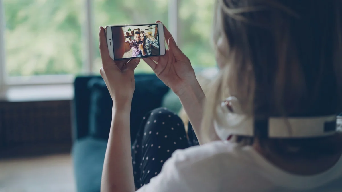 A young woman sitting in a bright room, holding up a phone to watch a video