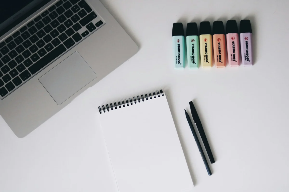 A laptop, a white notebook, two pens, and six pastel highlighters on a white desk