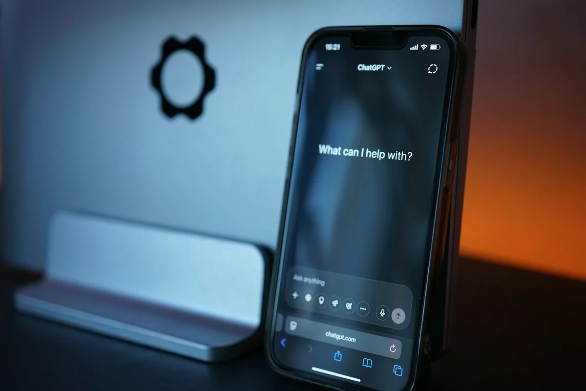 A phone open to chatgpt.com in a browser with the line What can I help with, propped up next to a laptop