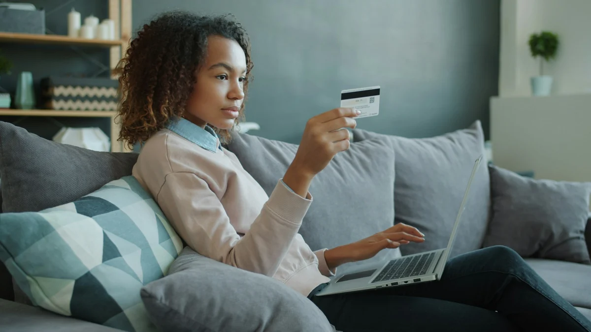 A young woman on a sofa holding a payment card and typing on a laptop