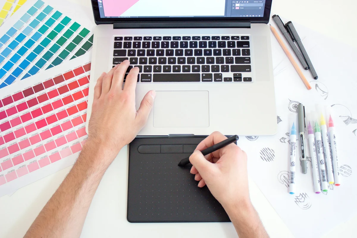 A designer using a drawing tablet next to a laptop, with color swatches and colored pens on a white desk