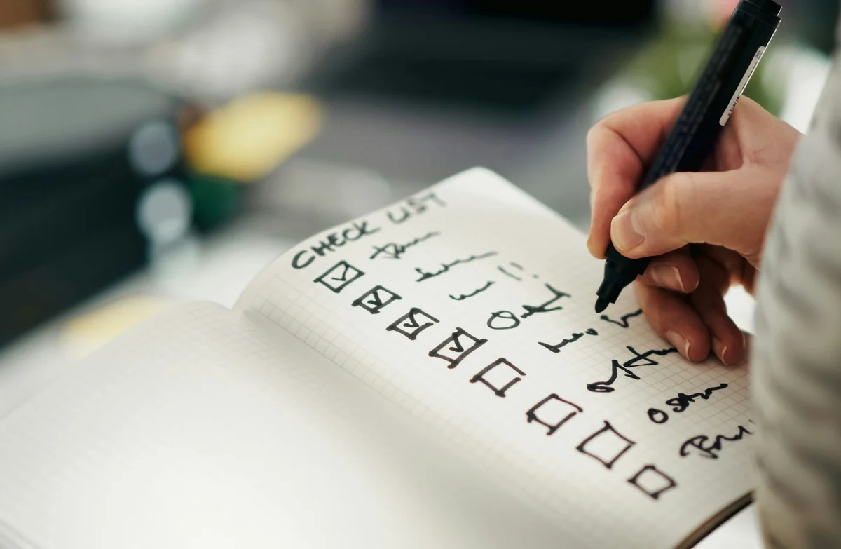 A hand holding a marker, checking boxes on a handwritten checklist in a notebook