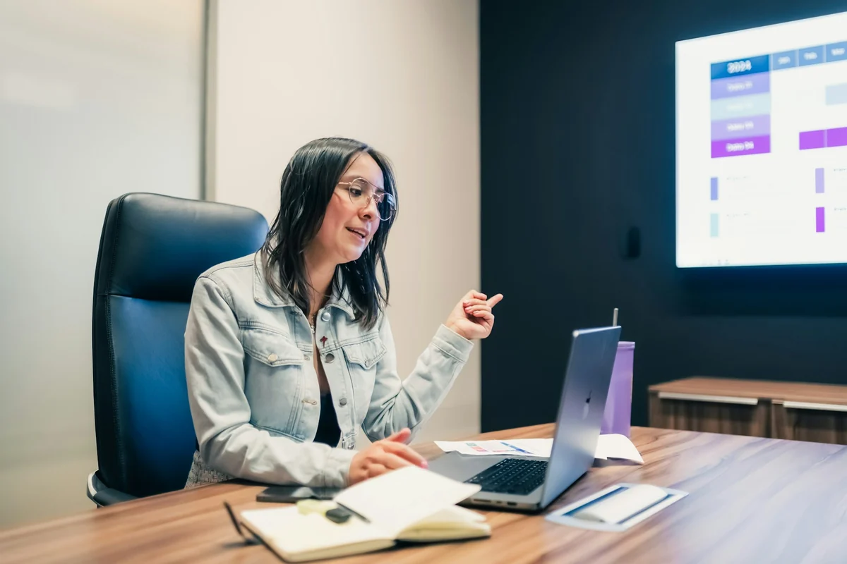 A woman sitting at a laptop giving a presentation, with a meeting room screen behind her showing a colorful slide