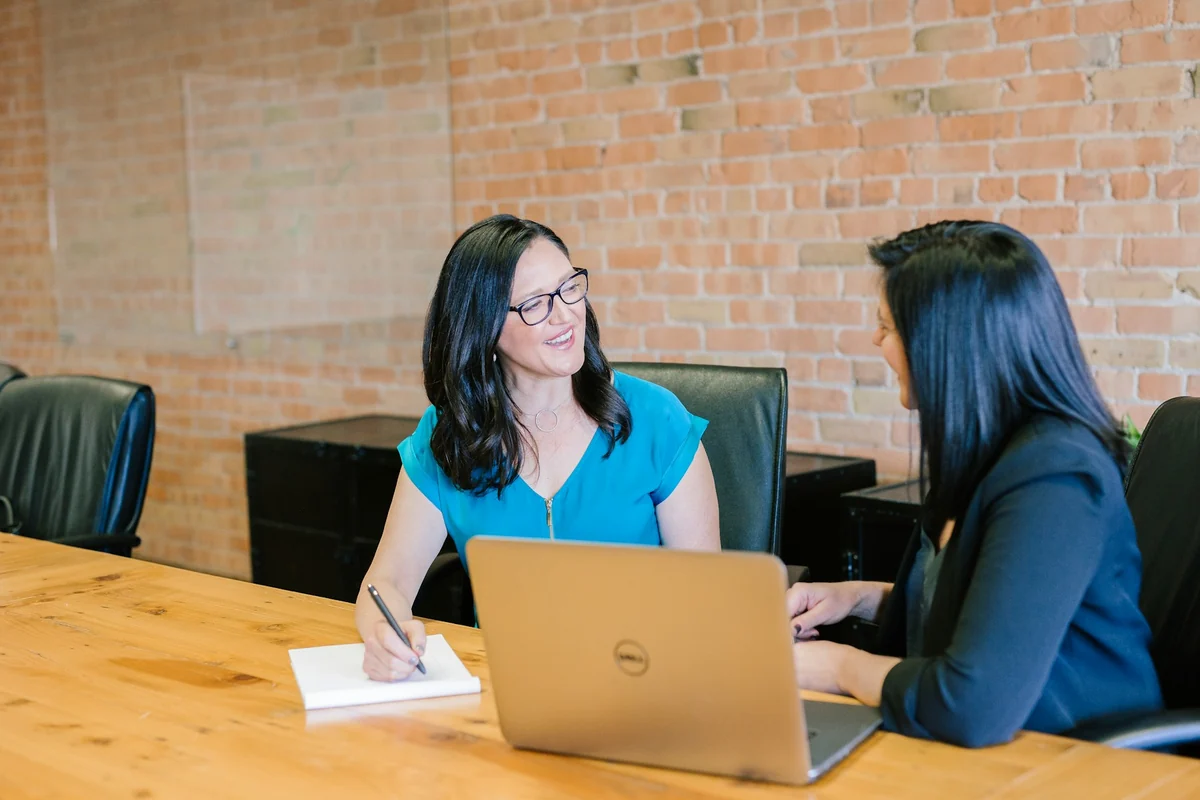 Two women sit at a wooden table discussing work, one taking notes next to a laptop