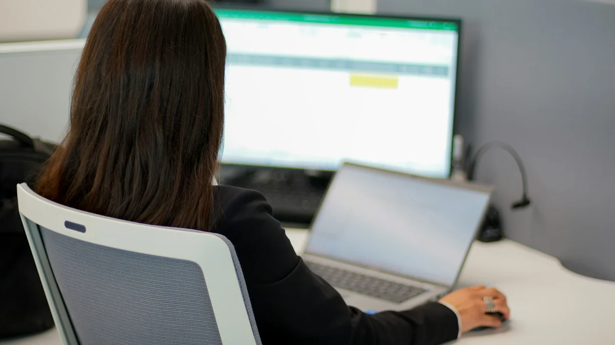 Office worker sitting at a large monitor showing a spreadsheet, with a laptop beside it
