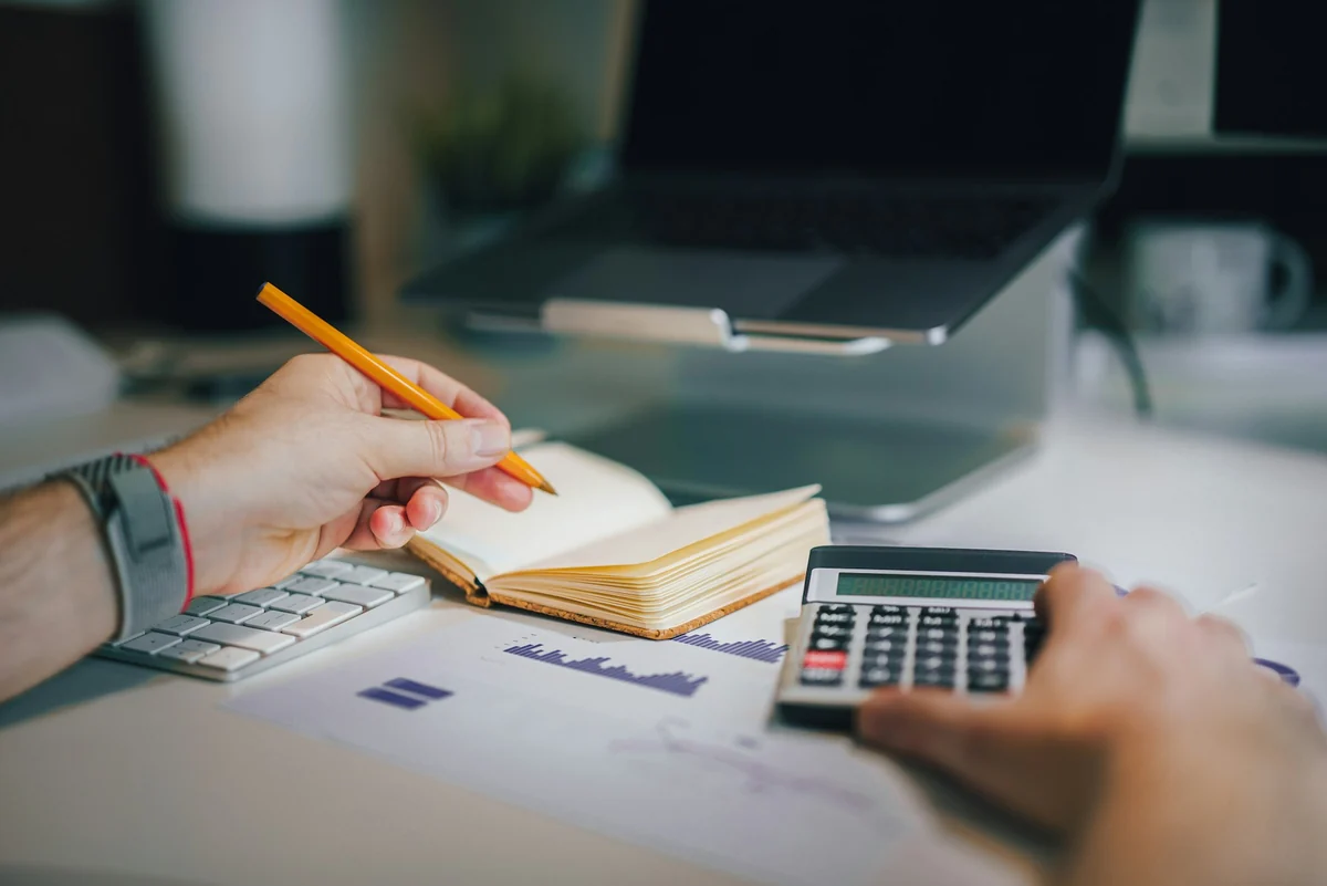 Two hands holding a pencil and tapping a pocket calculator, checking figures next to a notebook and a laptop