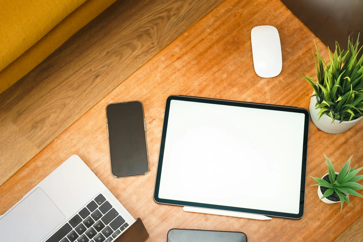 A laptop, a tablet, and a phone side by side on a wooden table with a green potted plant