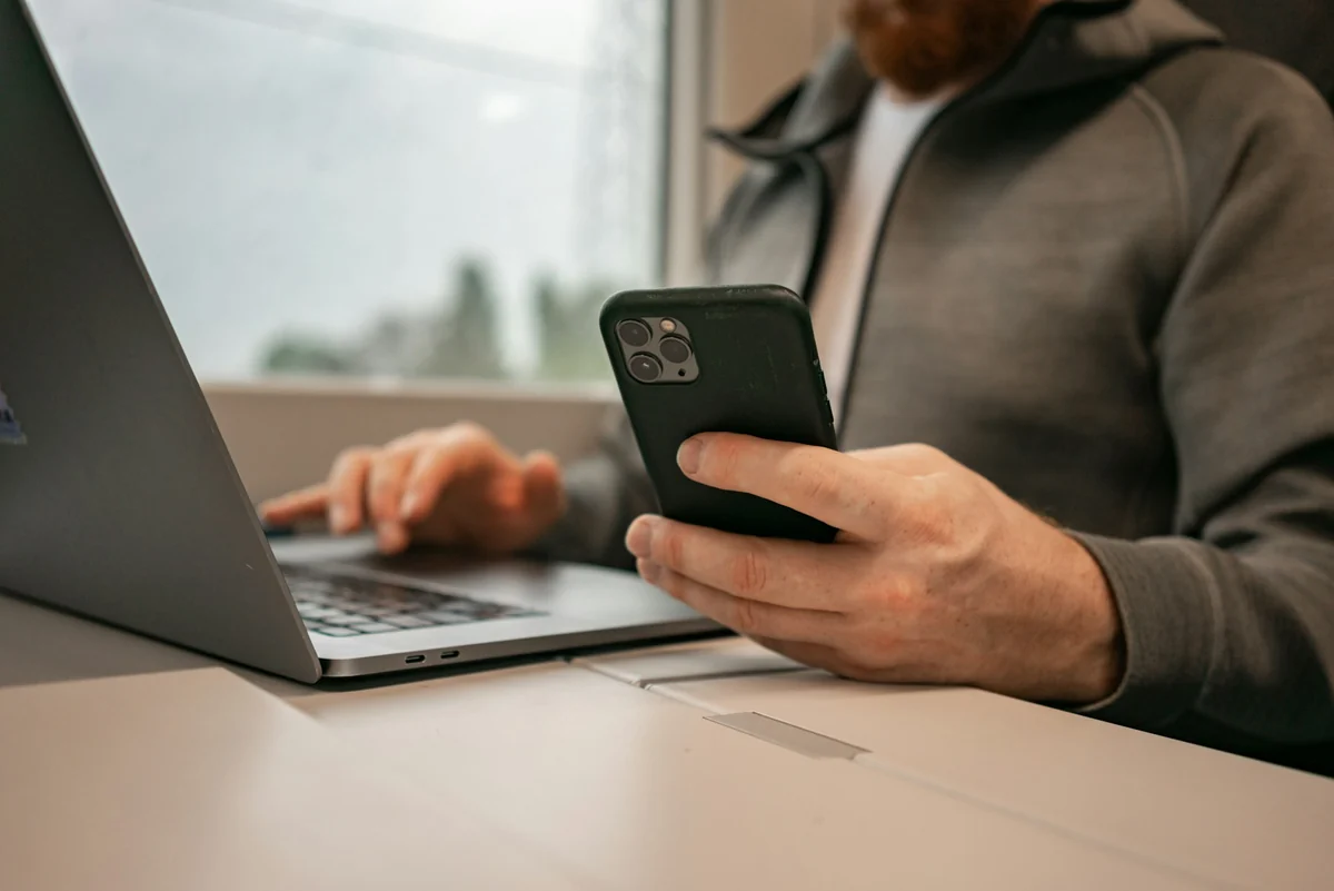 Người đàn ông cầm iPhone trong tay trái, tay phải gõ laptop cạnh cửa sổ
