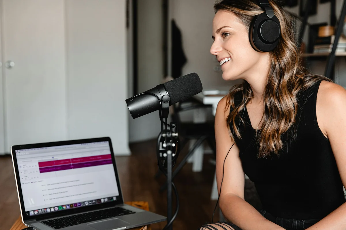 A woman wearing headphones speaks into a microphone next to a laptop running recording software