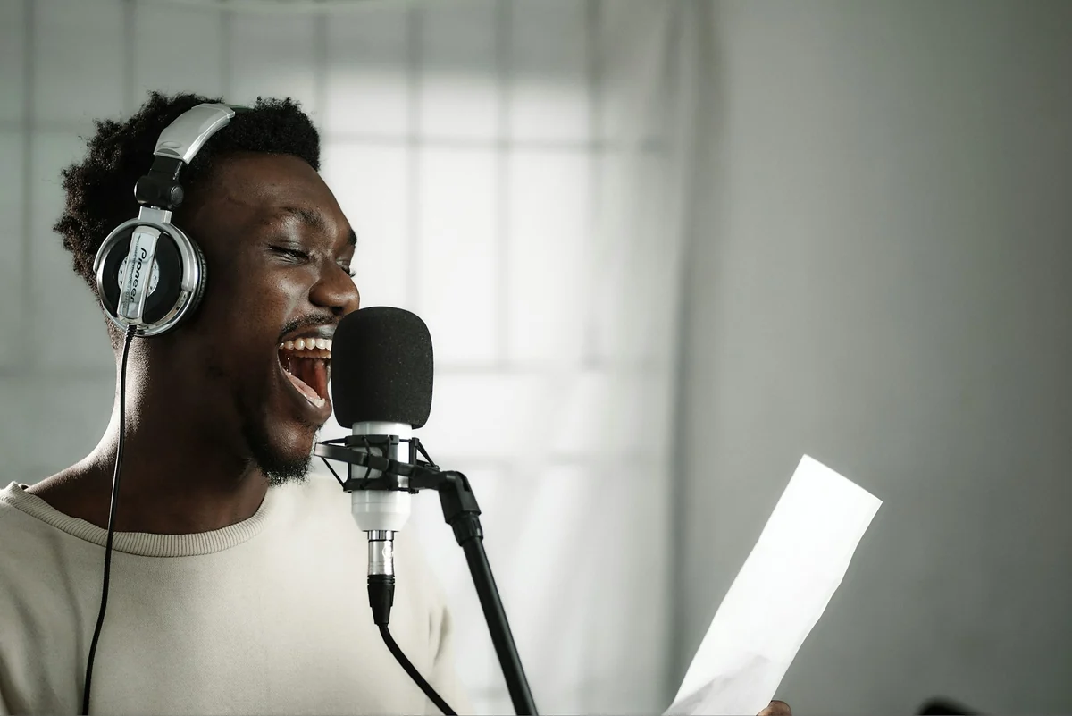 A man wearing headphones, holding a script and reading into a recording microphone