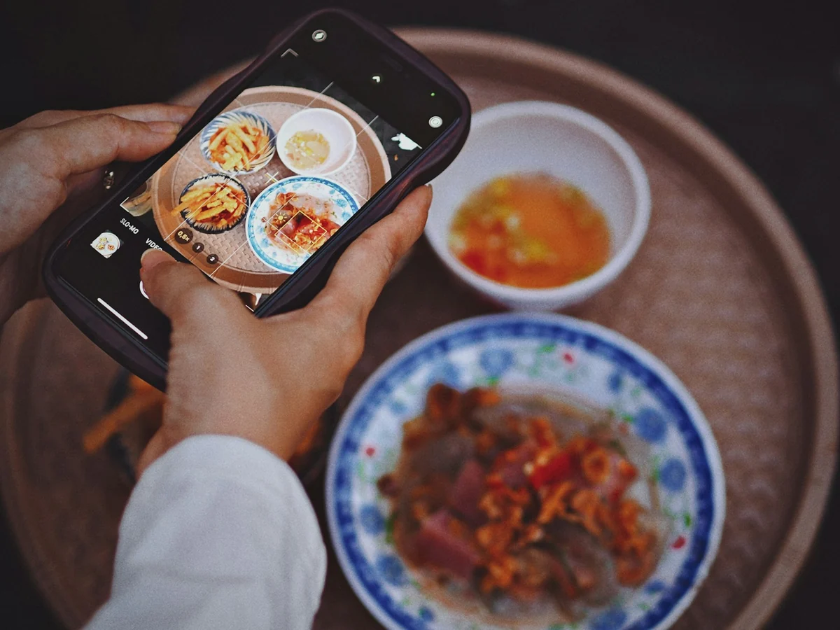 Two hands holding a phone to photograph a plate of food and a small bowl of dipping sauce on a tray