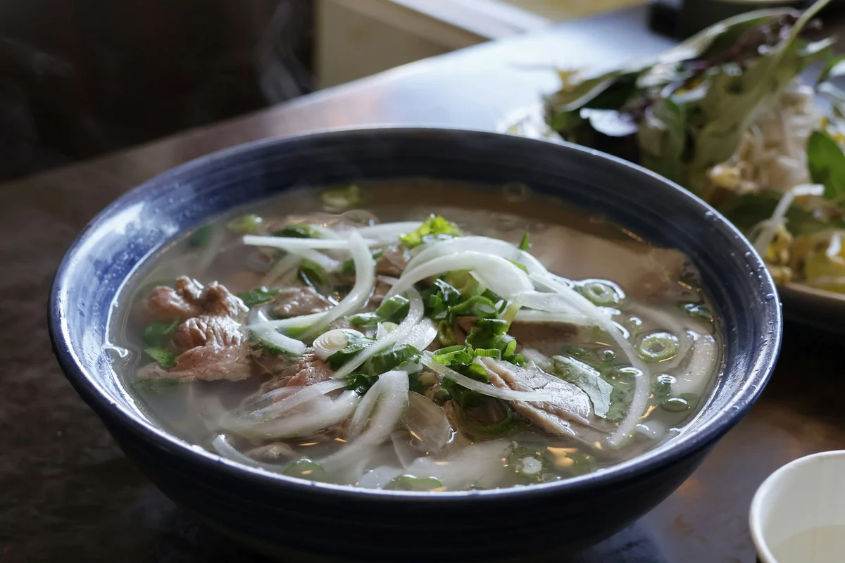 Bowl of beef pho with onions and green onions in a dark blue glazed bowl, shot at an angle from above