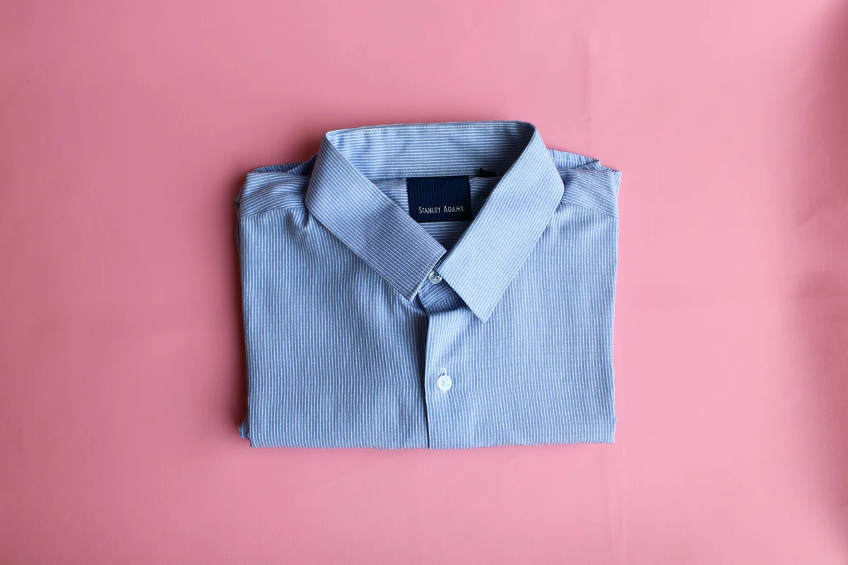 A folded blue striped shirt on a plain pink fabric background, the kind of product photo that works as the first image