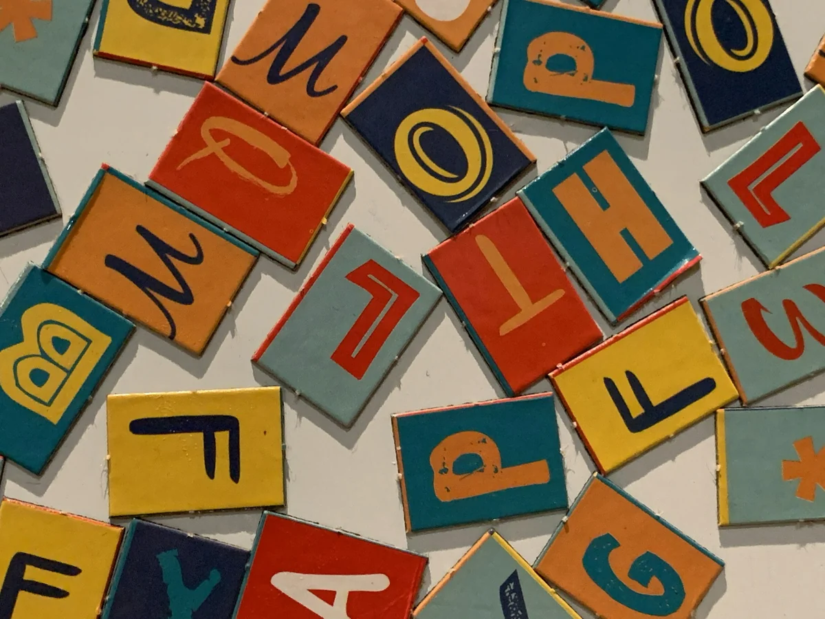 Many painted wooden letters in red, blue, and yellow piled on top of each other, each in a different typeface
