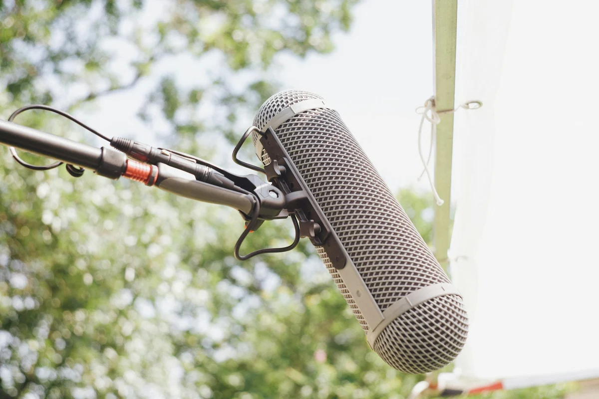 A shotgun microphone with a windscreen on a boom pole, set up outdoors to record background sound