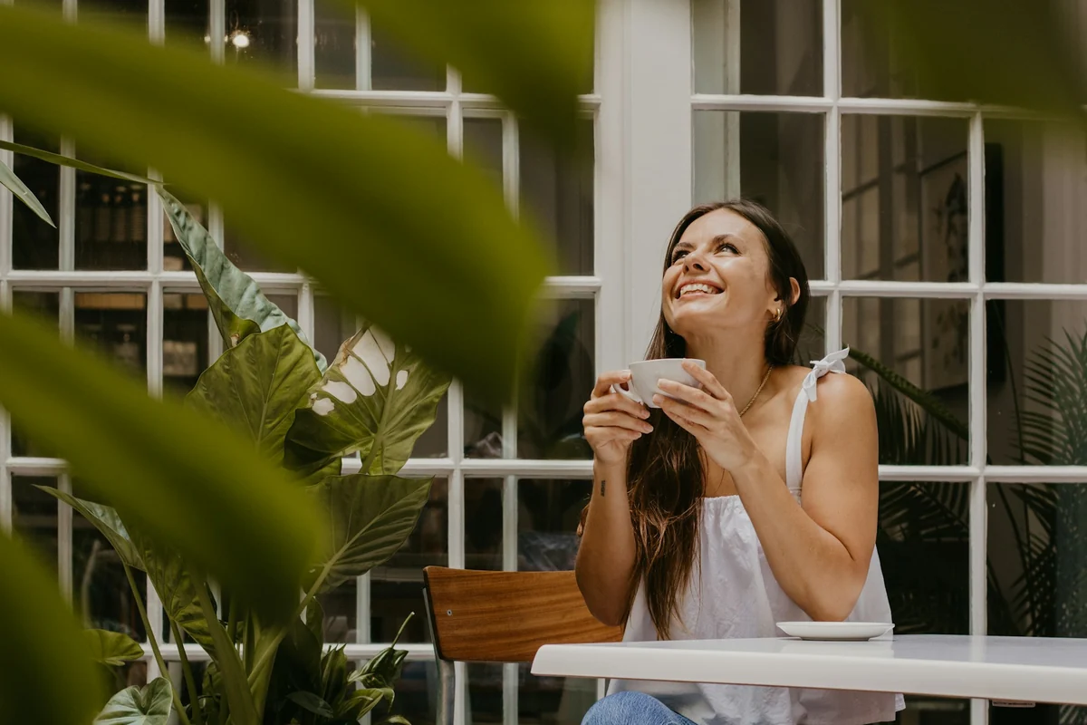 A young woman holds a coffee cup and smiles by a window in a cafe full of plants