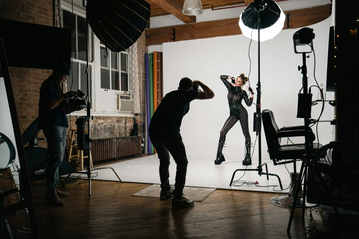 A photographer leans in to shoot a model in a studio with lights and a white backdrop
