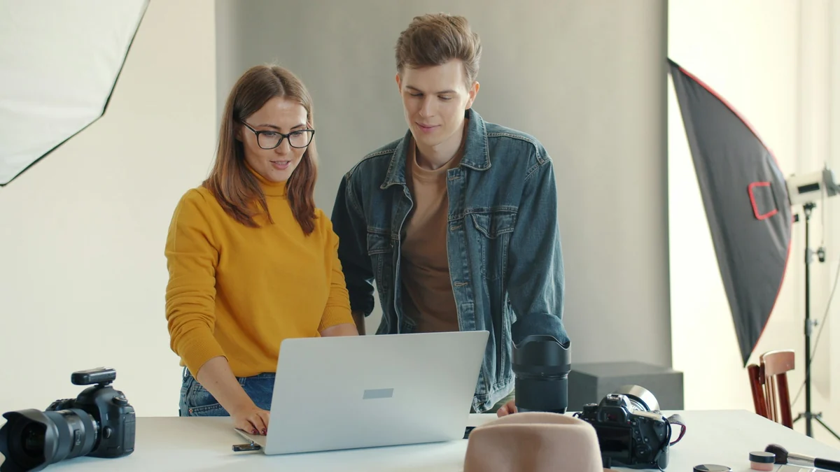 Two people in a studio review photos together on a laptop, with a camera beside it