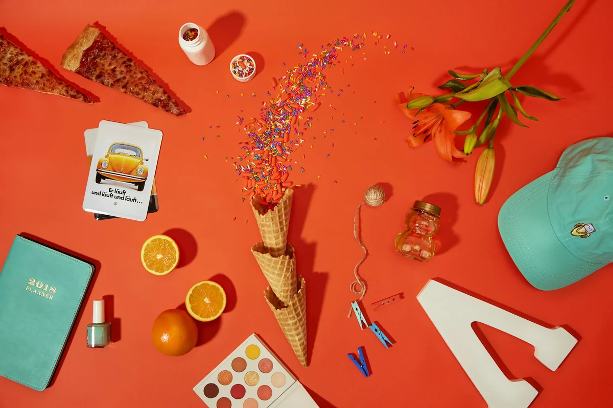 A flat lay of items on a red background: a notebook, an ice cream cone, oranges, flowers, and a baseball cap