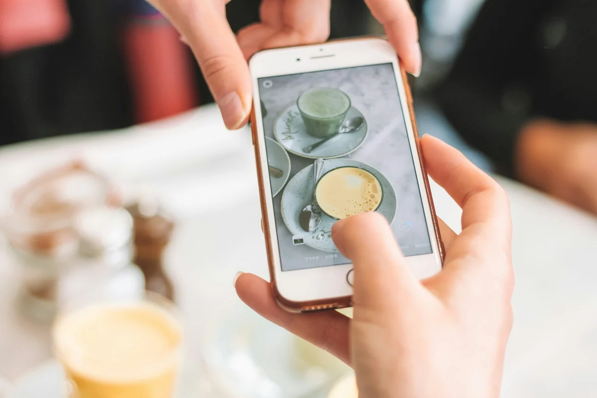 Two hands holding an iPhone to photograph two drinks on a white table