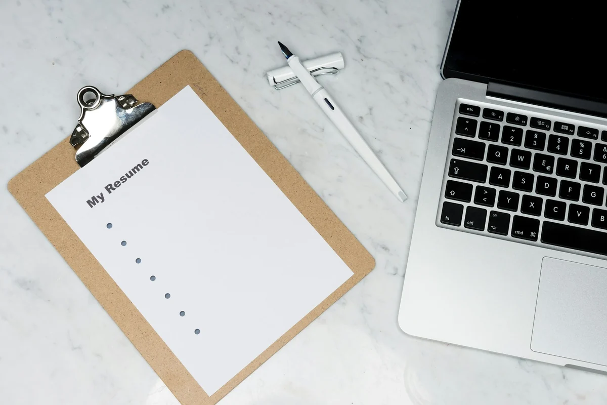 A folder holding a sheet titled My Resume next to a laptop and a pen on a stone desk