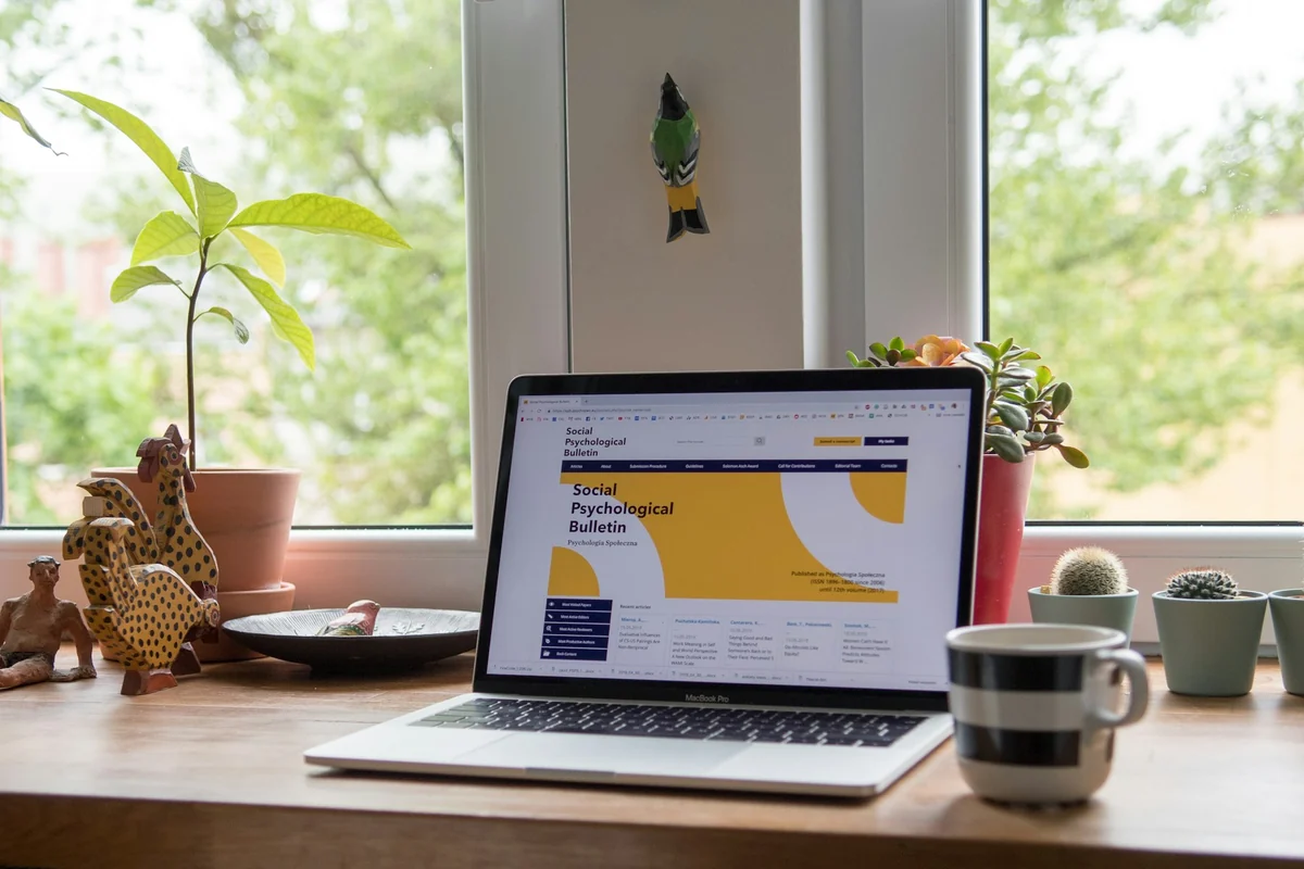 Laptop with an open browser on a desk by a window, next to a potted plant and a ceramic mug