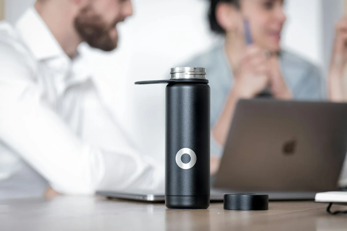 A black insulated bottle on an office desk, with two people talking over a laptop in the background