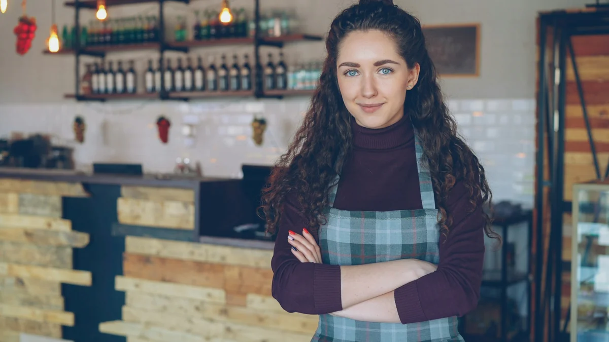 A woman in a checked apron stands with her arms crossed, smiling in front of a coffee shop counter