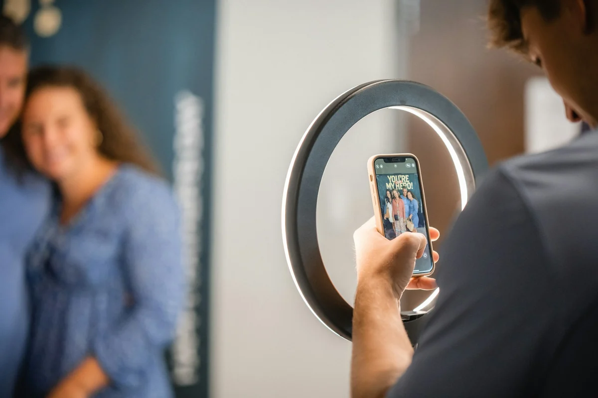 A person films a vertical video through a ring light, with a smiling woman standing in front