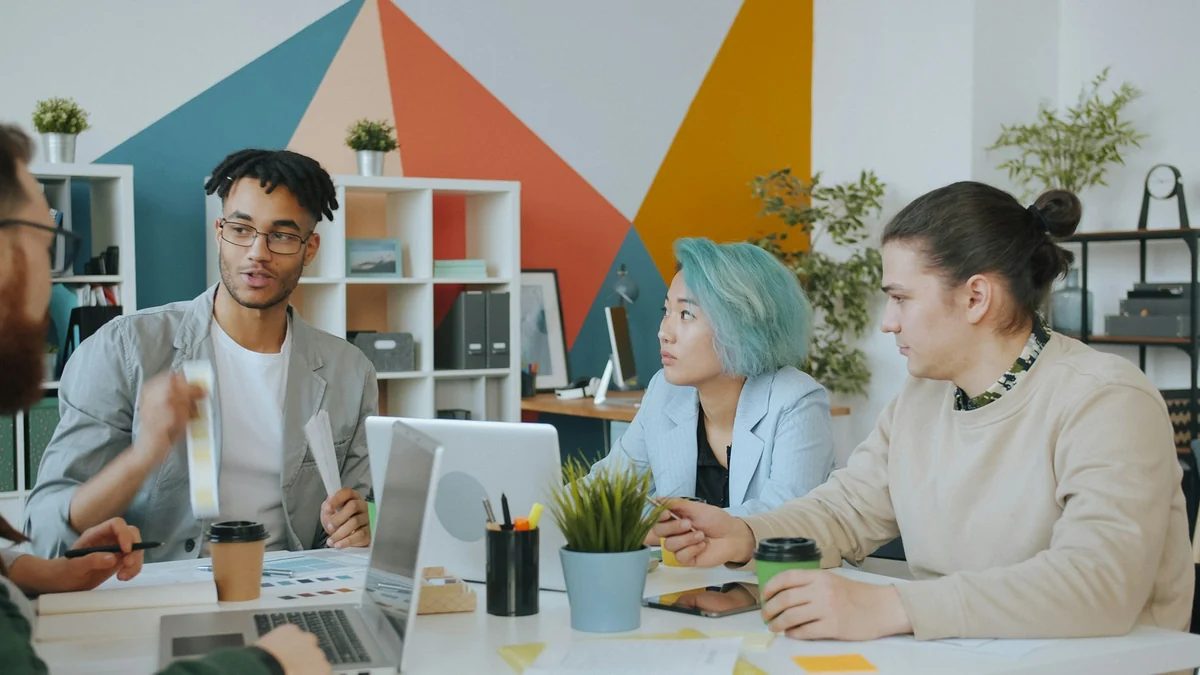 Four people around a meeting table in an office with multicolored painted walls, one of them presenting