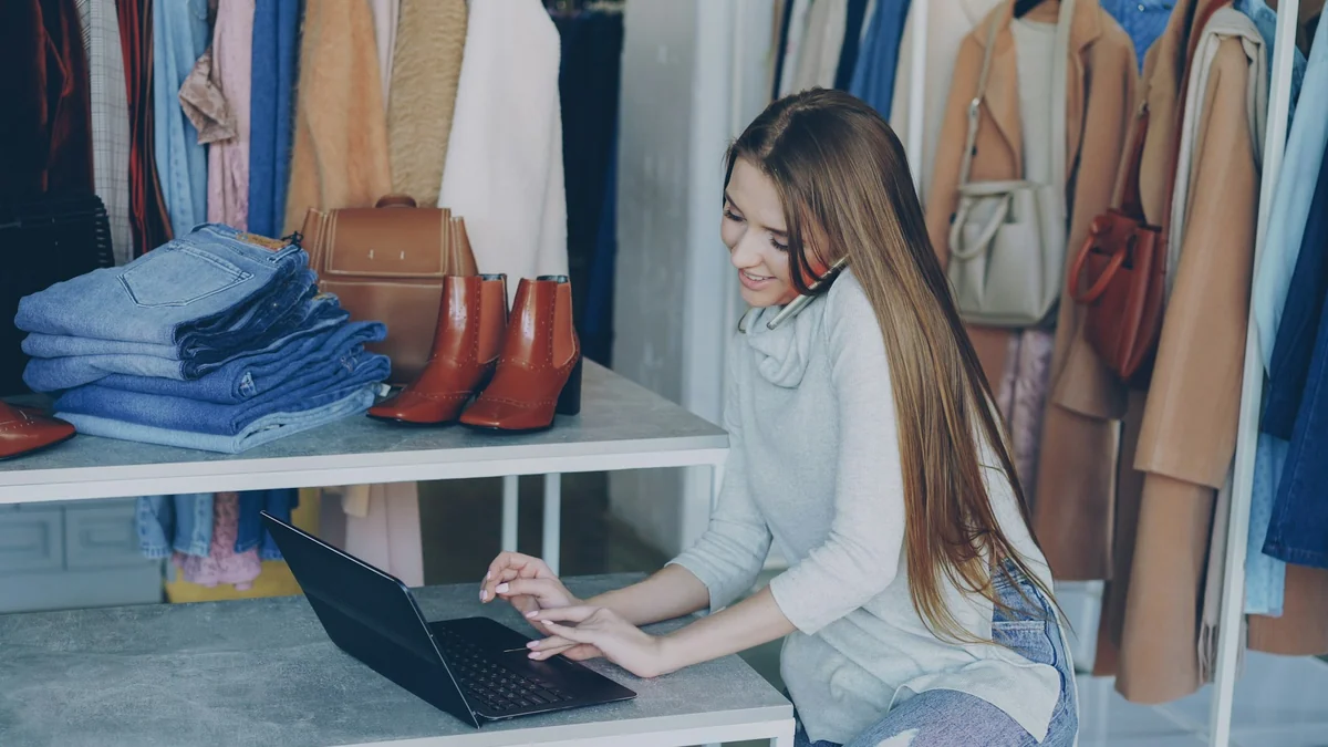 Clothing shop owner talking on the phone and typing on a laptop between racks of clothes