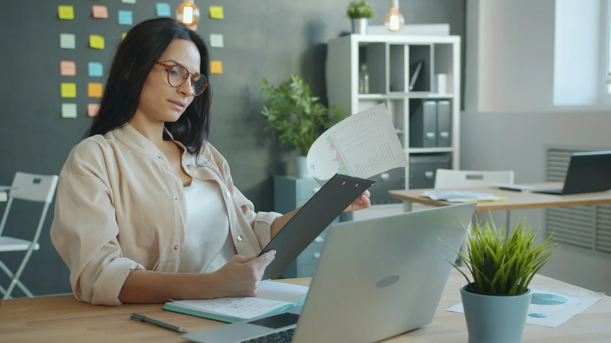 A woman in glasses holding a printed data sheet and comparing it with a laptop in an office