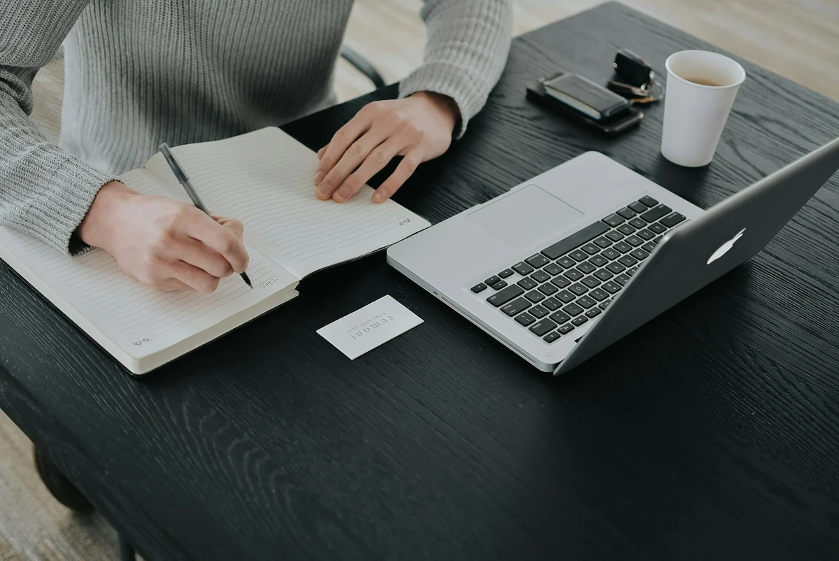 A person in a gray sweater taking notes in a notebook next to a laptop and a cup of coffee