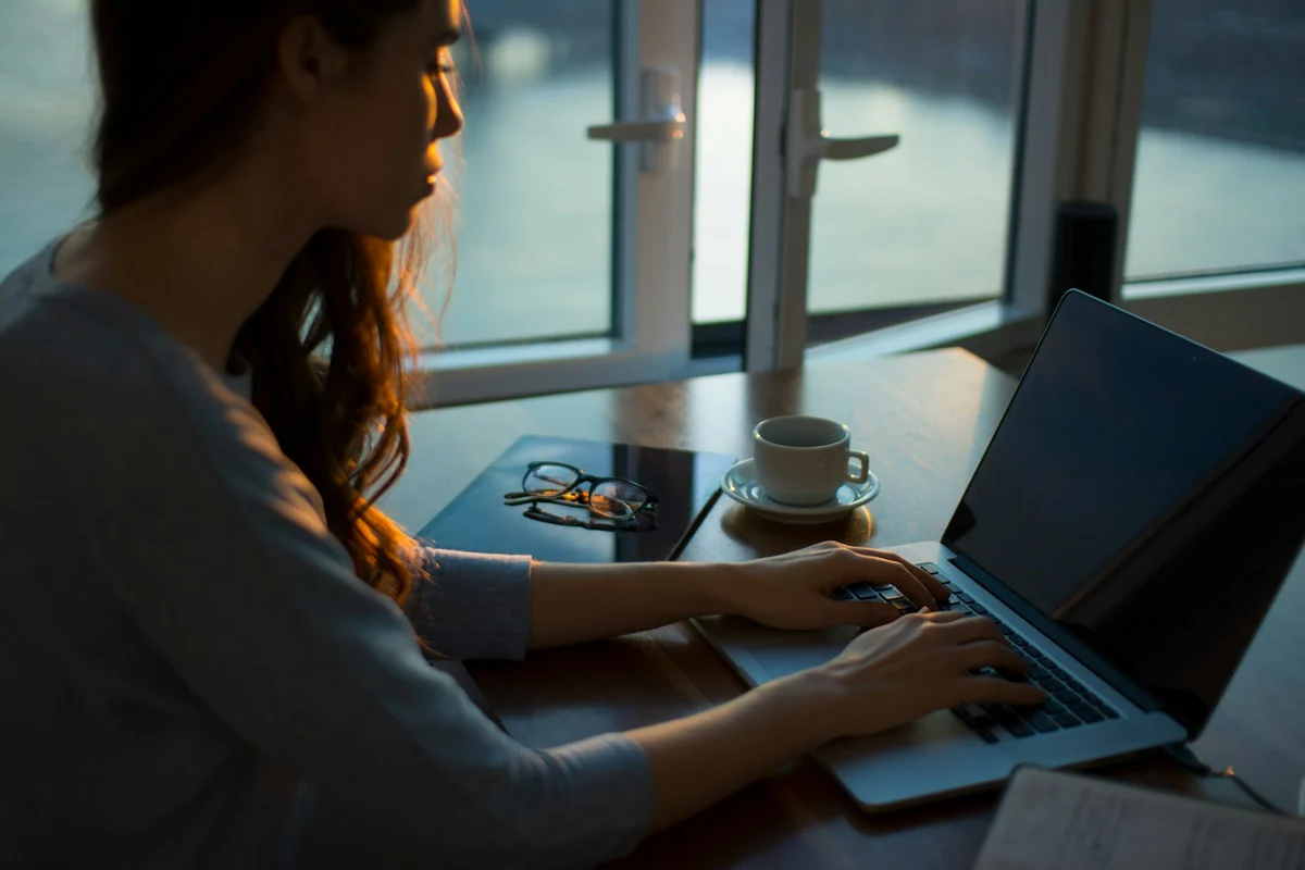 A woman by a window typing on a laptop, next to a cup of coffee and a pair of glasses on the table