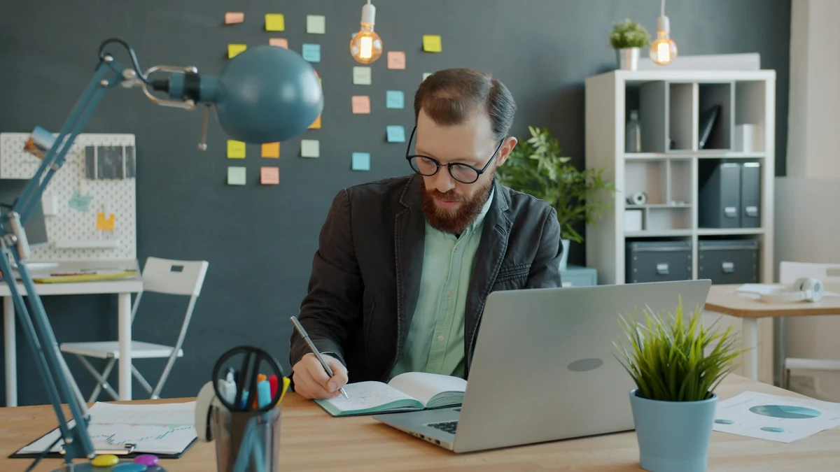 A man in glasses writing in a notebook next to a laptop in an office with sticky notes on the wall