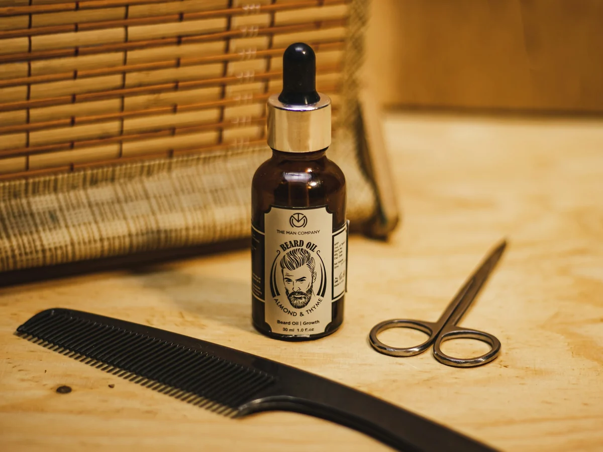 A bottle of beard oil on a wooden table next to a comb, scissors, and a bamboo blind
