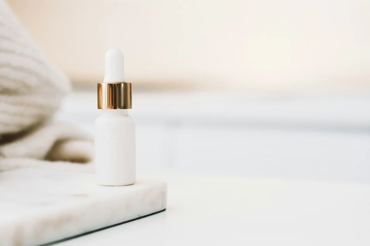 A white serum bottle with a gold cap on a marble tray, in front of a bright plain background
