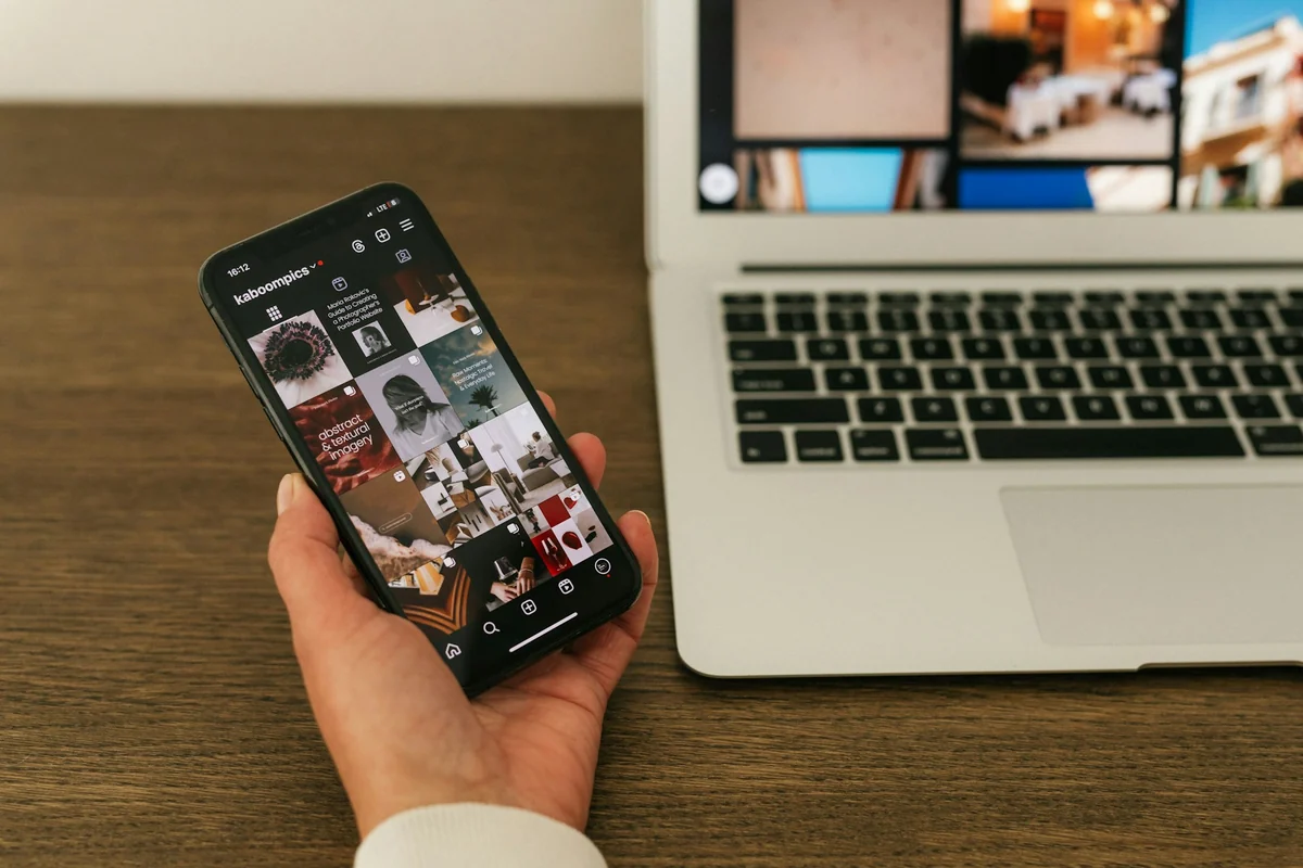 A hand holding a phone with a photo gallery open, next to a laptop on a wooden desk