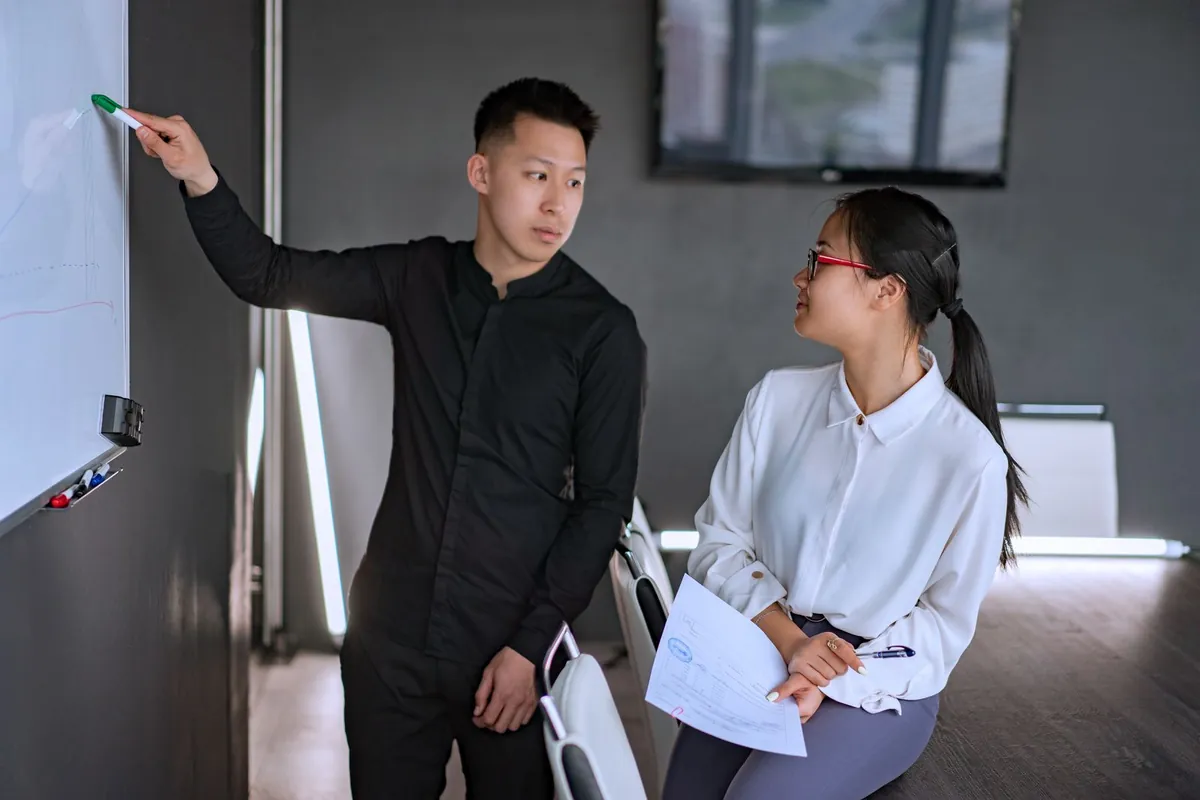 A manager points at a whiteboard, briefing a colleague who holds a printout